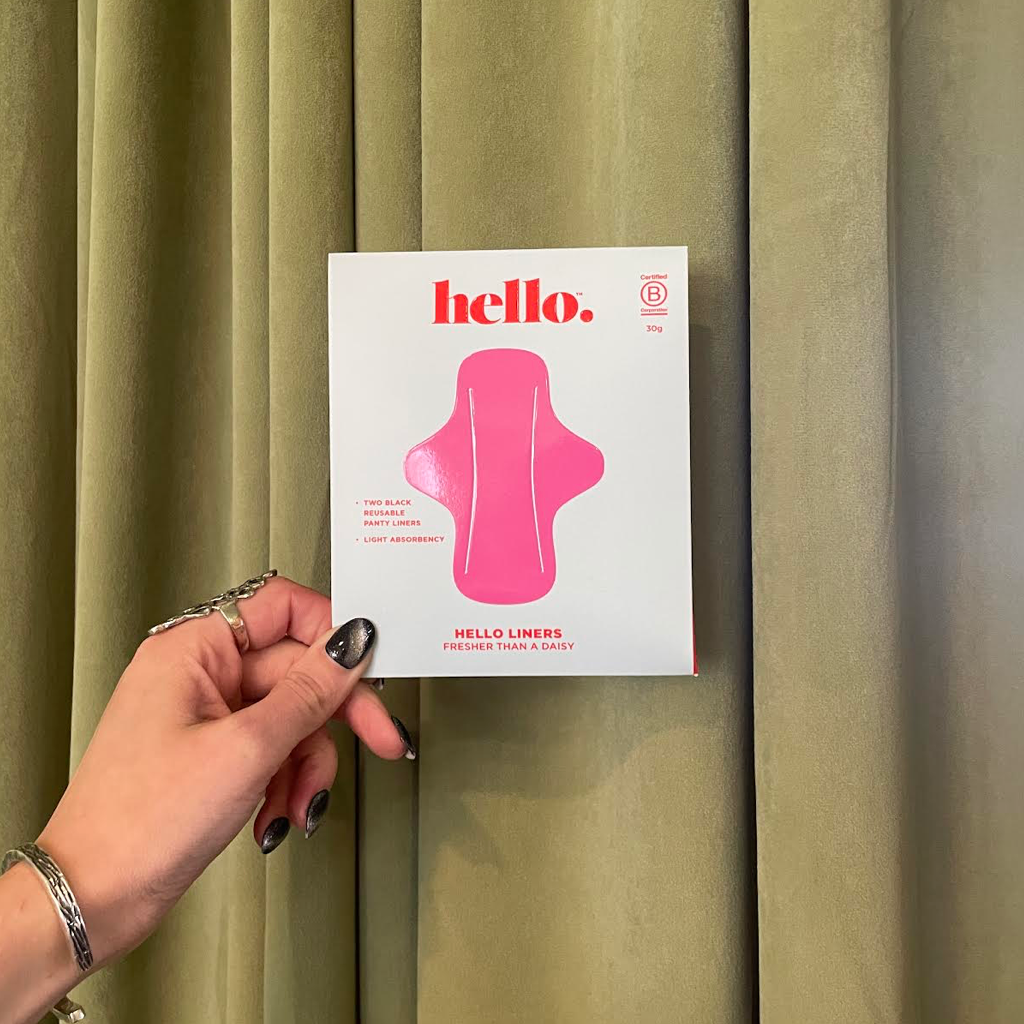 A hand with silver rings holds a box of Hello Liners 2 Pack- Fresher Than A Daisy ultra thin panty liners against olive green curtains. The white box displays a pink pad illustration and red text promoting quick-wick technology and the brand name.