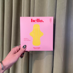 A hand with silver rings and dark nail polish holds a pink Hello Pads 2 Pack- Ride or Die box containing reusable period pads, in front of greenish-grey curtains.
