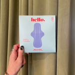 A hand with black-painted nails holds a box of Hello Pads 2 Pack- Overnight Success for heavy flow in front of beige curtains. The light blue box features a purple pad illustration and product info in red and white text.