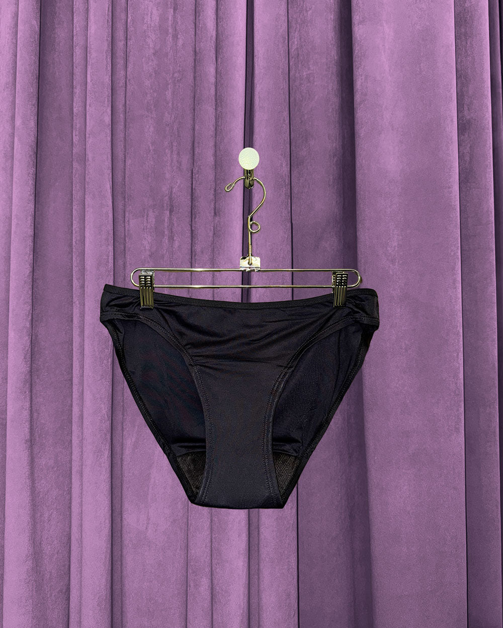 A pair of Period Swim Bottoms - Bikini Cut in black hangs on a metallic hanger, set against purple velvet curtains.