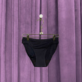A pair of Period Swim Bottoms - Bikini Cut in black hangs from a gold hanger against purple velvet curtains.