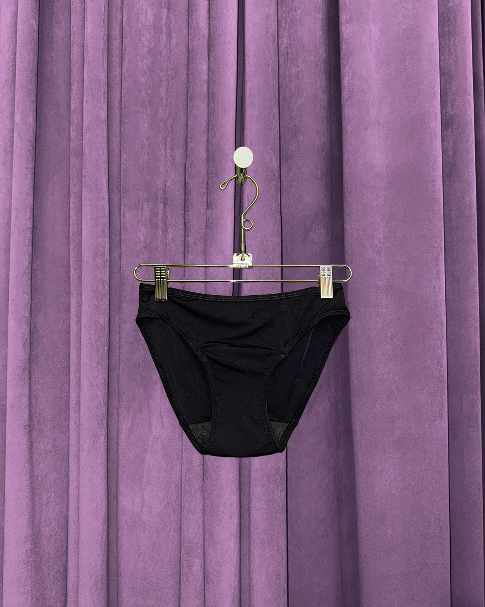 A pair of Period Swim Bottoms - Bikini Cut in black is displayed on a hanger in front of closed, pleated purple curtains.