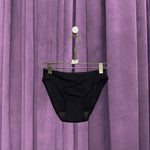 A pair of Period Swim Bottoms - Bikini Cut in black is displayed on a hanger in front of closed, pleated purple curtains.