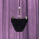 A pair of Period Swim Bottoms - Bikini Cut in black is displayed on a hanger in front of closed, pleated purple curtains.