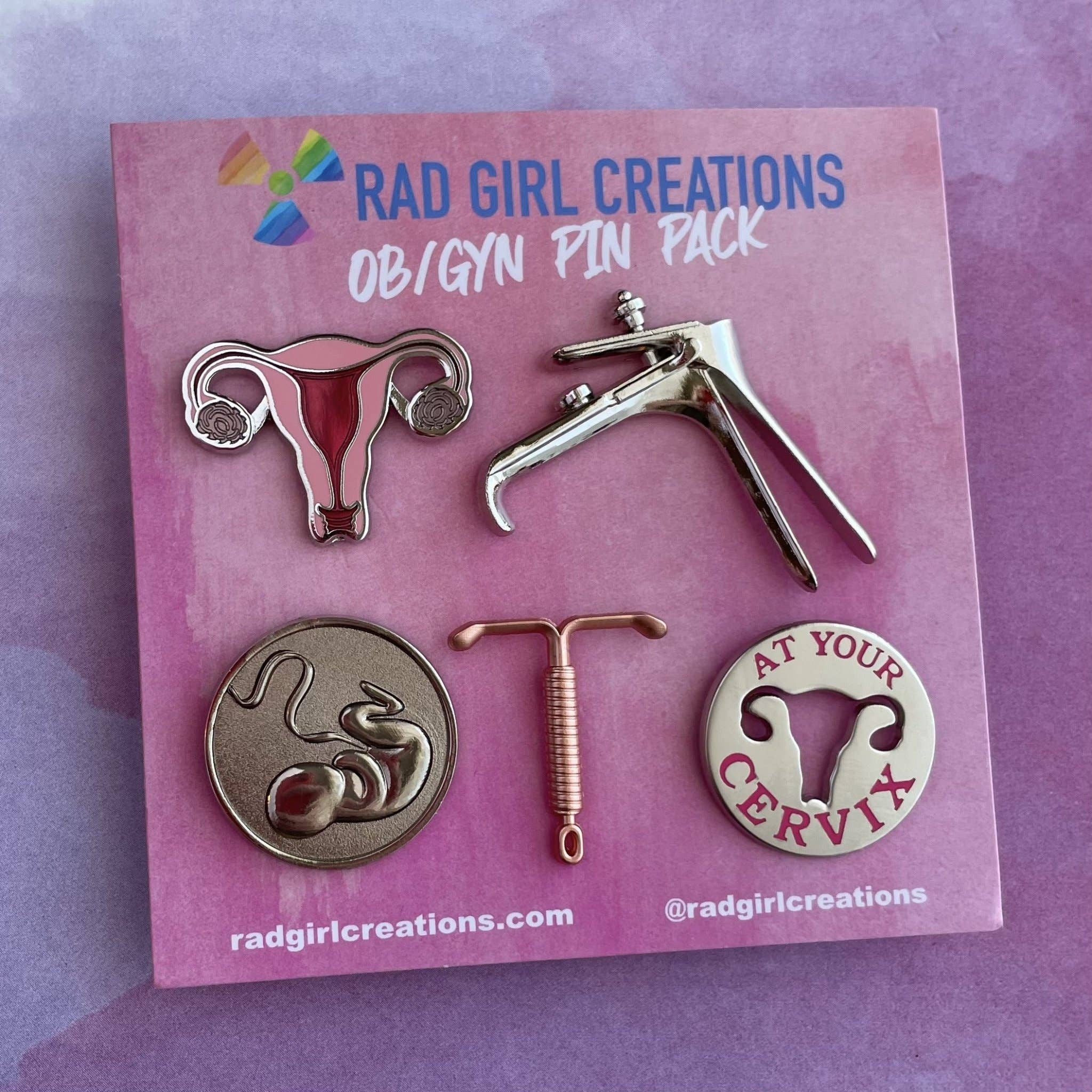 The OB/GYN Pin Pack includes five pins on a pink card: a uterus anatomy pin, speculum, fetus, IUD, and a round “AT YOUR CERVIX” pin with uterus artwork.