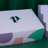 The Build Your Period Kit Bundle, in a rectangular white box with colorful geometric shapes and a stylized p logo, is displayed next to a four-step white staircase against a green background.