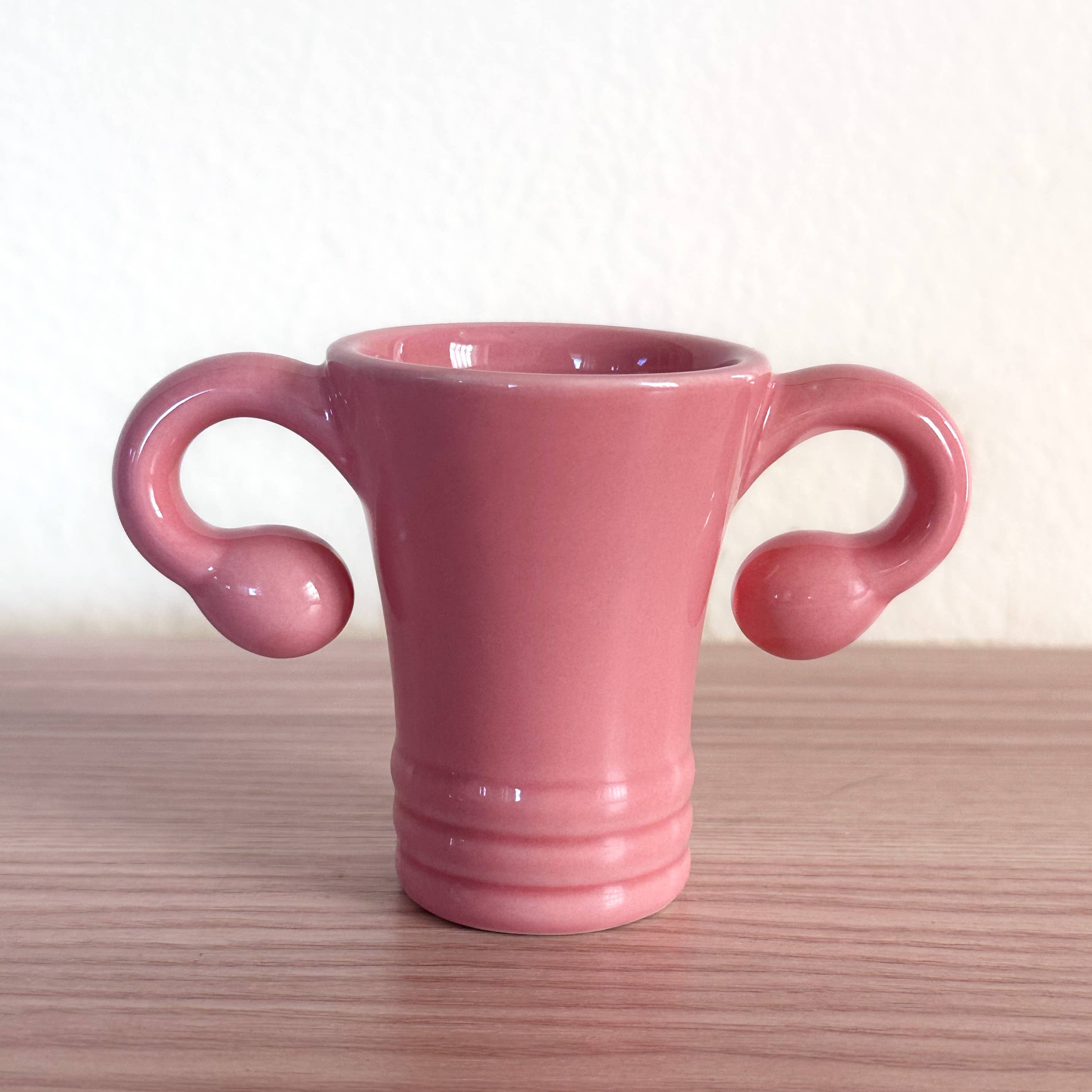 The Shooterus - Uterus Shot Glass is a pink ceramic cup shaped like a uterus, complete with fallopian tube handles—a playful novelty perfect for celebrating milestones like menopause or hysterectomy with humor.
