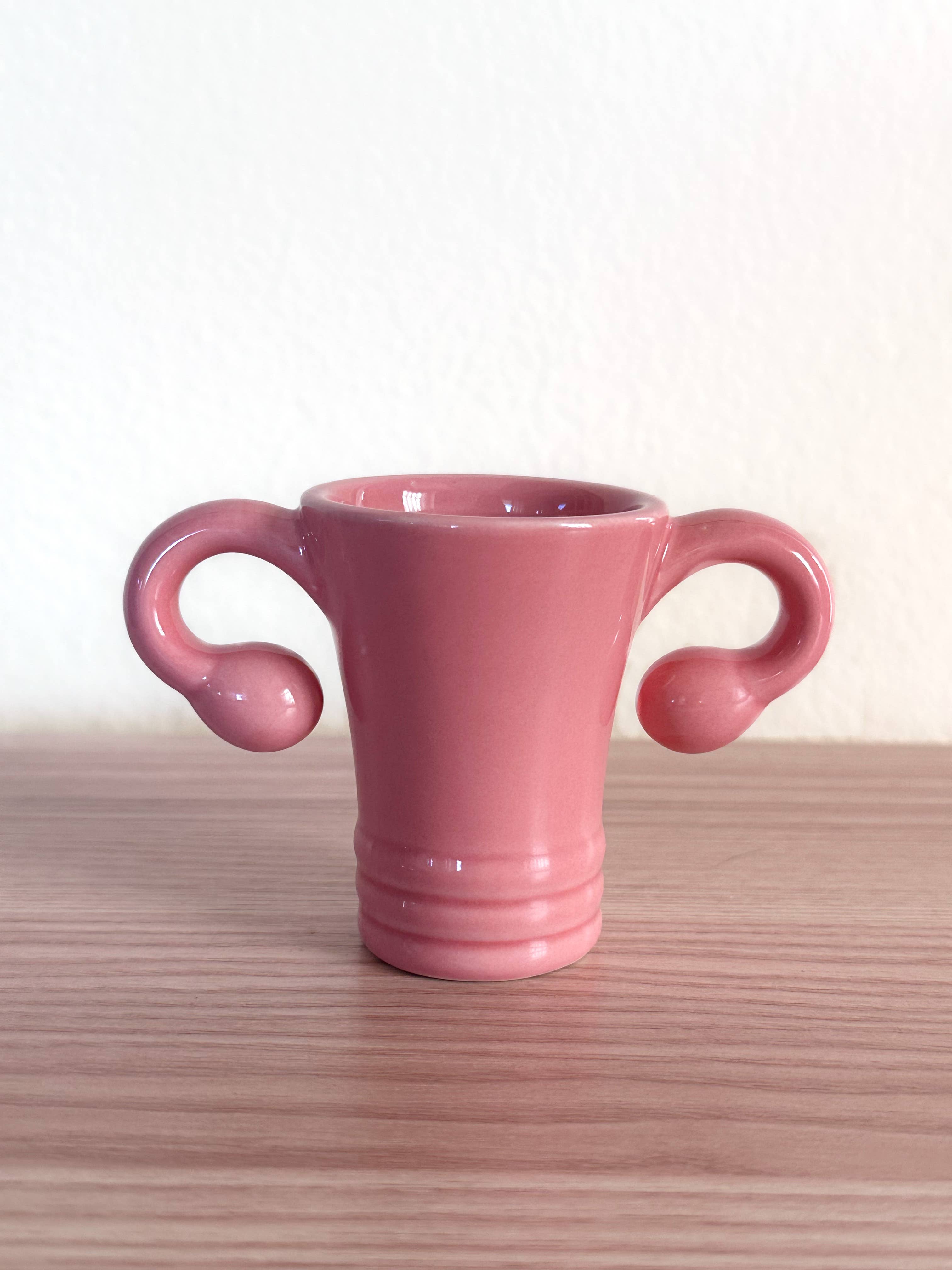 The Shooterus - Uterus Shot Glass is a pink ceramic cup shaped like a uterus, complete with fallopian tube handles—a playful novelty perfect for celebrating milestones like menopause or hysterectomy with humor.
