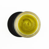 A top-down view of an open jar of Vital "V" Wild Yam Salve, a smooth greenish-yellow balm ideal as a natural intimate moisturizer or for vaginal dryness relief, set on a white background with a black circular shadow beneath the jar.