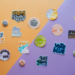 The "My Body My Choice" sticker, along with other colorful feminist and empowerment pins and stickers featuring slogans like “Strong Female Lead” and “Smash the Patriarchy,” is displayed on a diagonally split purple and orange background.