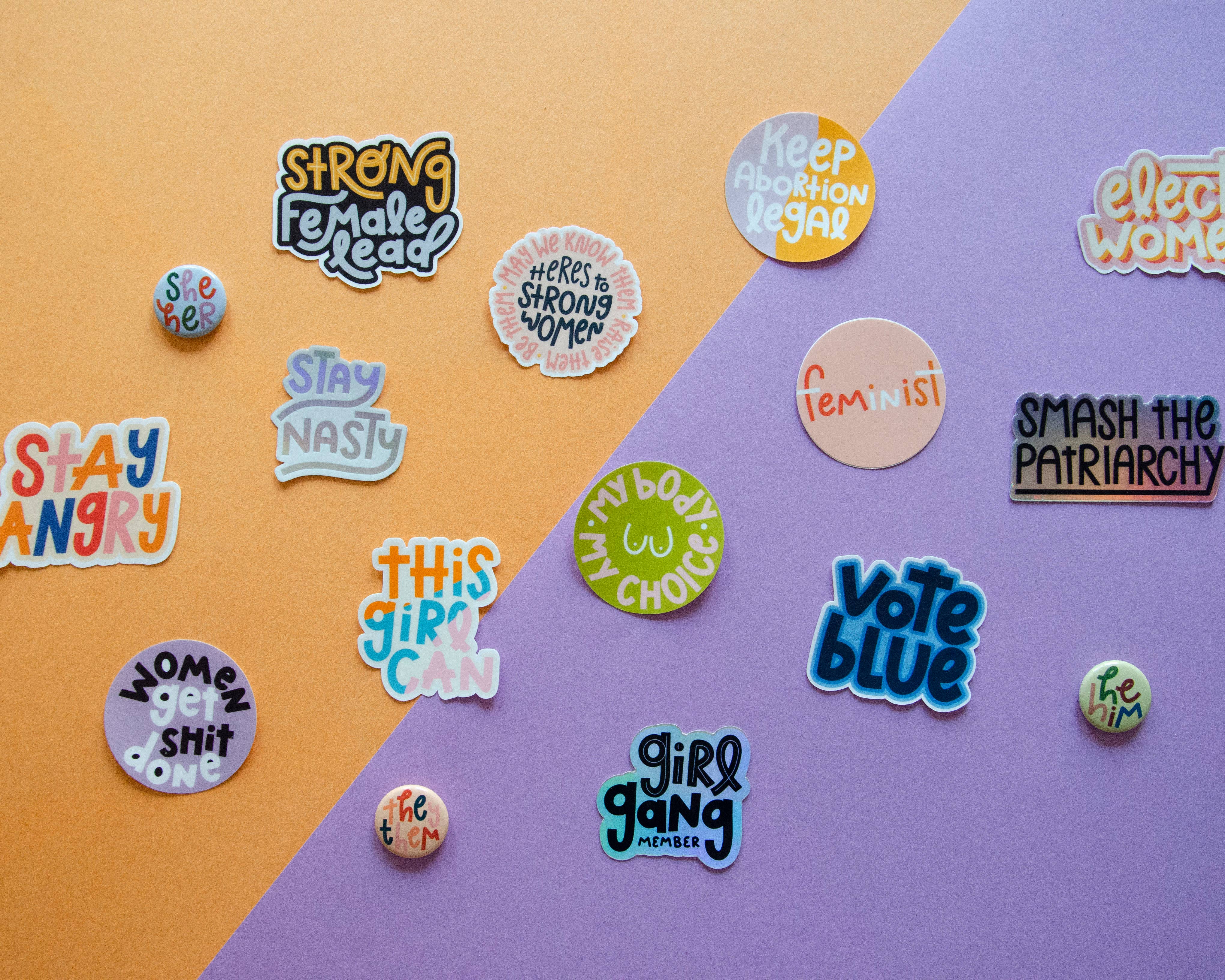 The "My Body My Choice" sticker, along with other colorful feminist and empowerment pins and stickers featuring slogans like “Strong Female Lead” and “Smash the Patriarchy,” is displayed on a diagonally split purple and orange background.