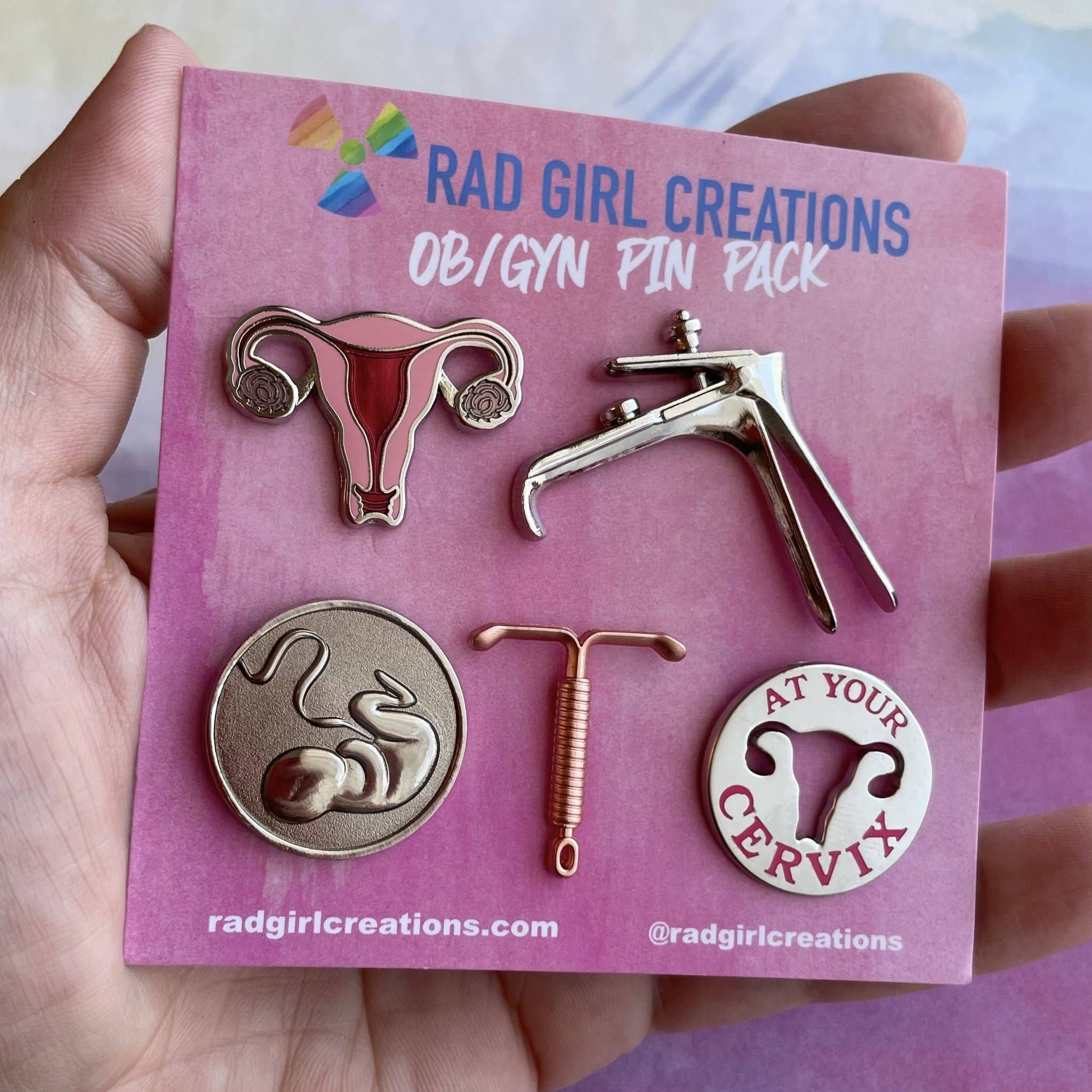 A hand holds the OB/GYN Pin Pack, which features five pins: a uterus, a speculum, a fetus in womb, an IUD, and a cervix anatomy pin with "AT YOUR CERVIX" text.