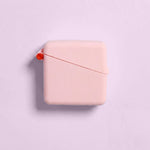A square, pale pink Hello Caddy measuring tape with rounded edges sits on a light pink background, resembling a compact carry case. A small red button and thin red measuring tape are partially visible at the top left corner.