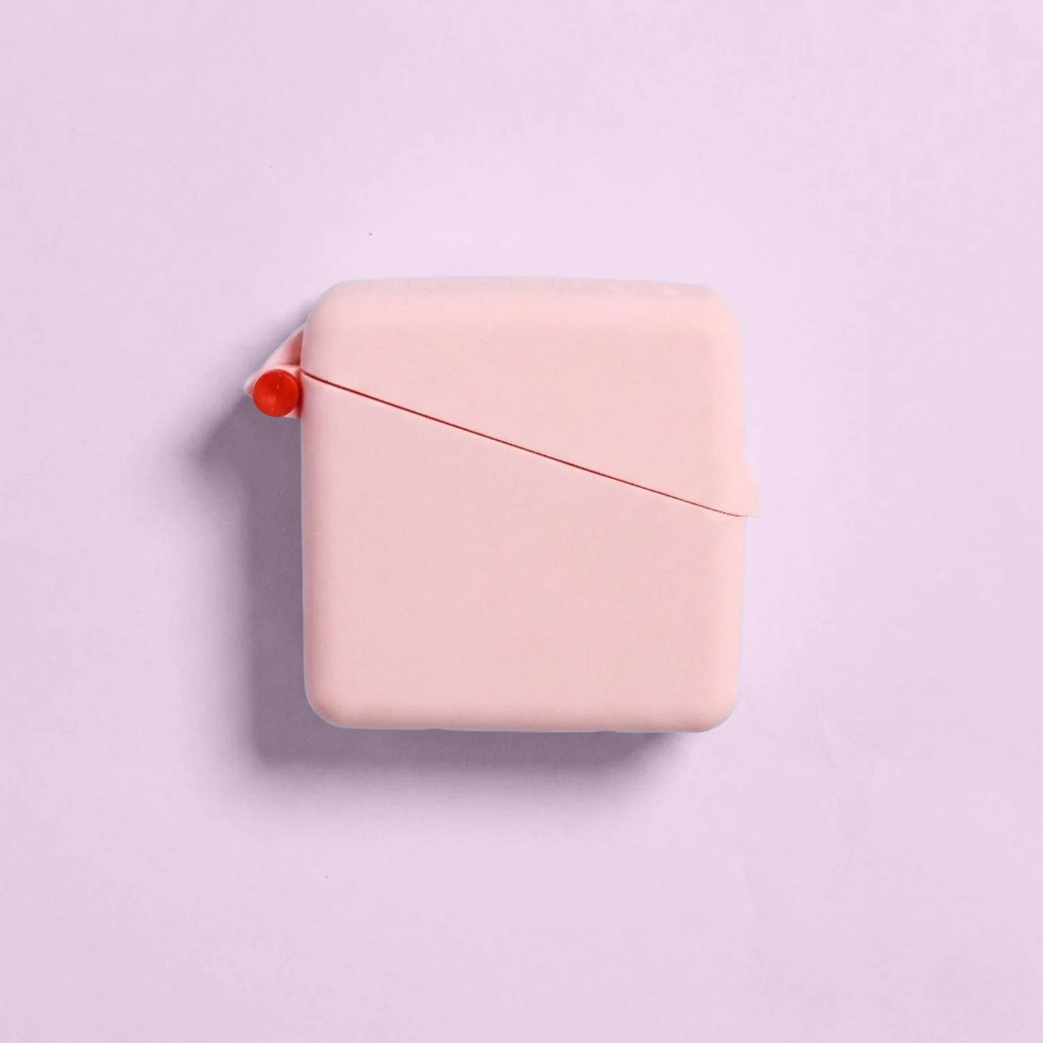 A square, pale pink Hello Caddy measuring tape with rounded edges sits on a light pink background, resembling a compact carry case. A small red button and thin red measuring tape are partially visible at the top left corner.