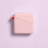 A square, pale pink Hello Caddy measuring tape with rounded edges sits on a light pink background, resembling a compact carry case. A small red button and thin red measuring tape are partially visible at the top left corner.