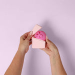 Two hands open the Hello Caddy, a small light pink container, revealing a round, pink item with face-like features inside, set against a light purple background.