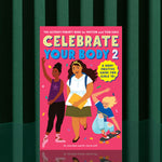 The book "Celebrate Your Body 2" stands upright against a green striped background, featuring three smiling, diverse girls—one dancing, one with books, and one skateboarding—on its cover.