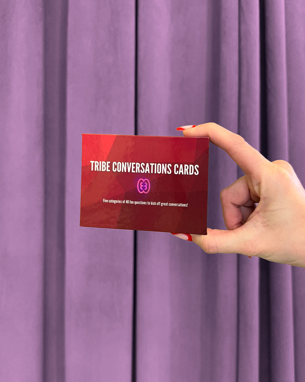 A hand with red-painted nails holds a box labeled Red Tribe Period Conversation Cards in front of purple curtains. The box features five categories with 41 fun questions—ideal for growing up discussions and great conversations!.