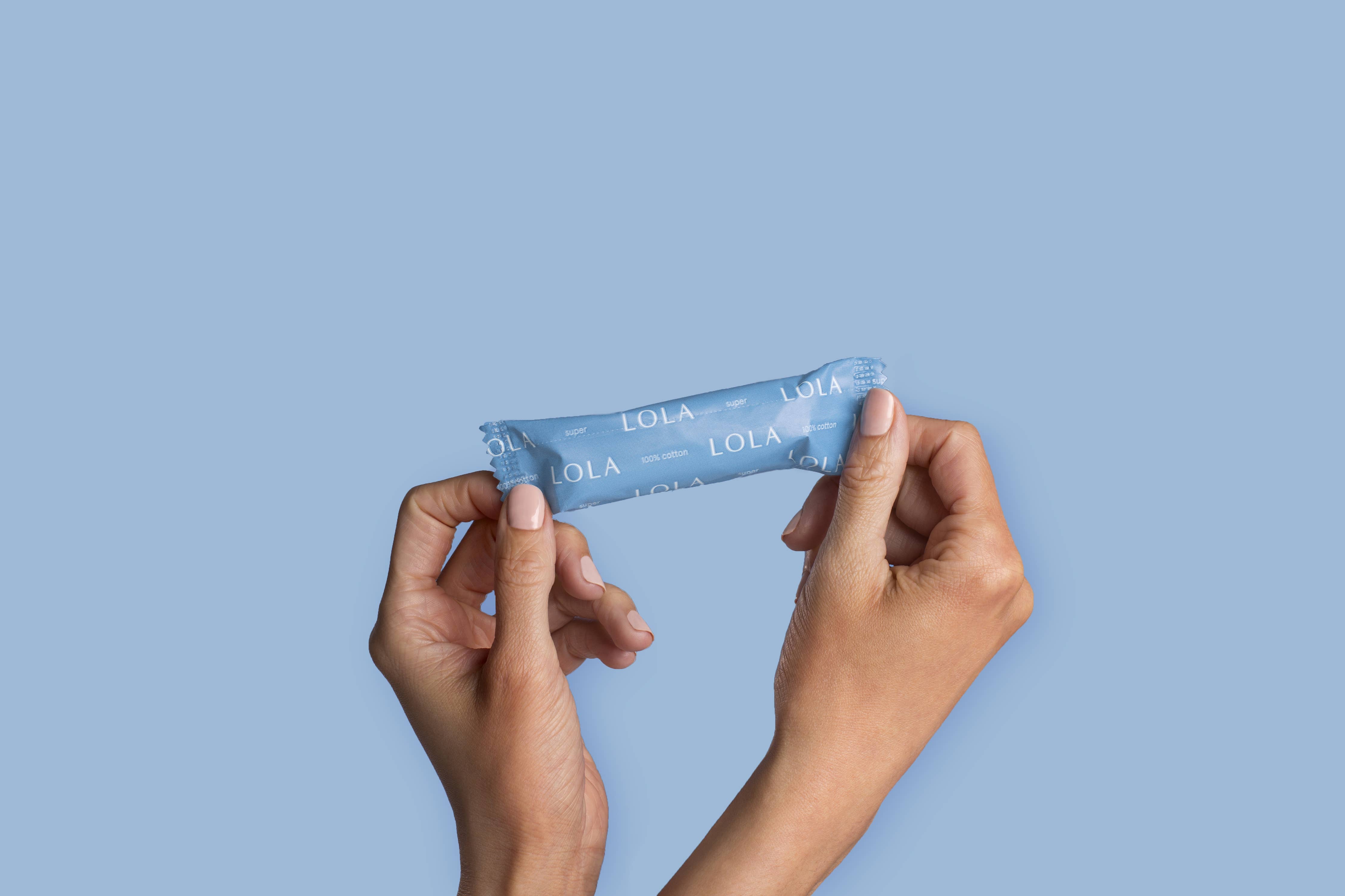 Two hands hold a box of Lola 20ct Super Tampons, featuring BPA-free applicators, against a solid light blue background.