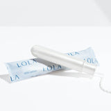 A Lola 20ct Light Tampon with a BPA-free applicator rests beside its light blue LOLA wrapper on a white surface, casting a soft shadow.