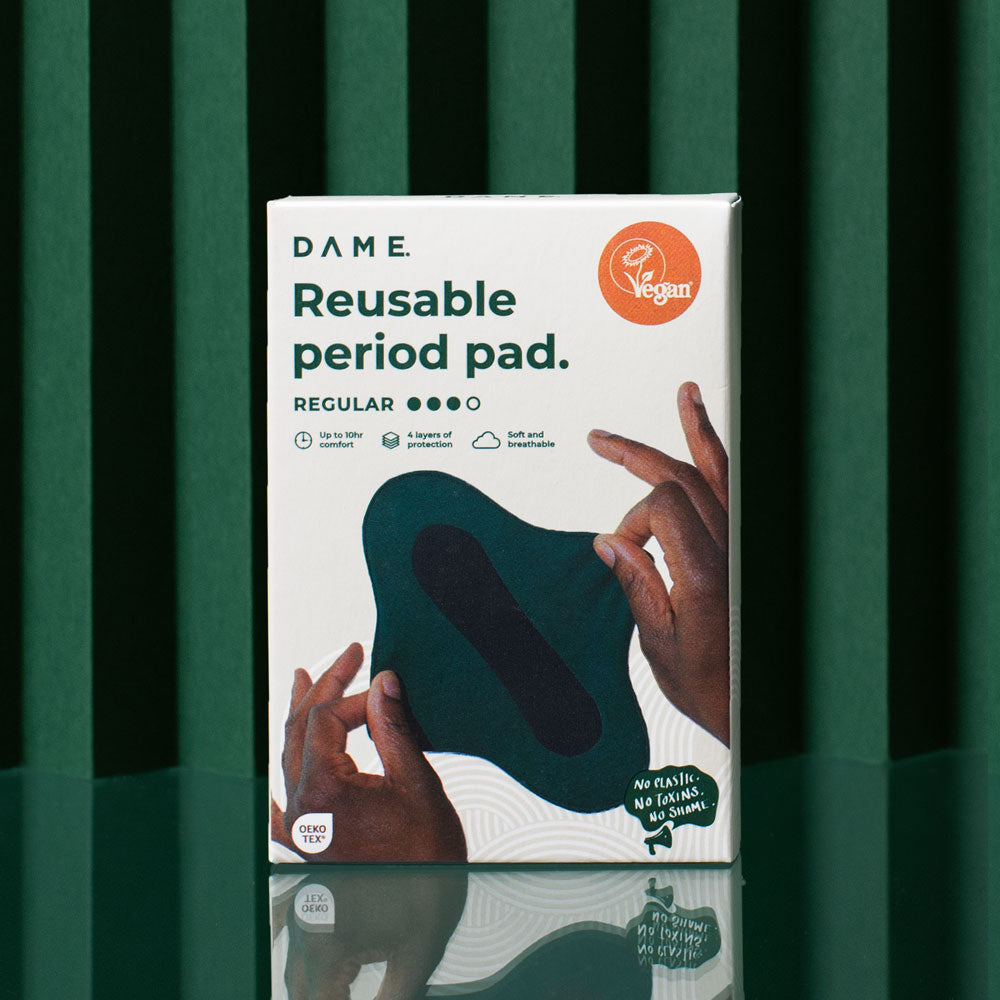 A box of DAME Cloth Pads is shown against a green backdrop. The packaging features a photo of the pad in two hands, highlights DAMEdry technology, and displays a “Vegan” certification badge.