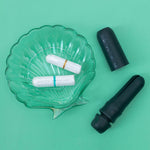 A glass seashell dish holds two tampons—one blue, one orange—beside two black DAME Reusable Tampon Applicators (one open, one closed), offering a hygienic, plastic-free period solution on a green background.