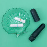 A glass seashell dish holds two tampons—one blue, one orange—beside two black DAME Reusable Tampon Applicators (one open, one closed), offering a hygienic, plastic-free period solution on a green background.