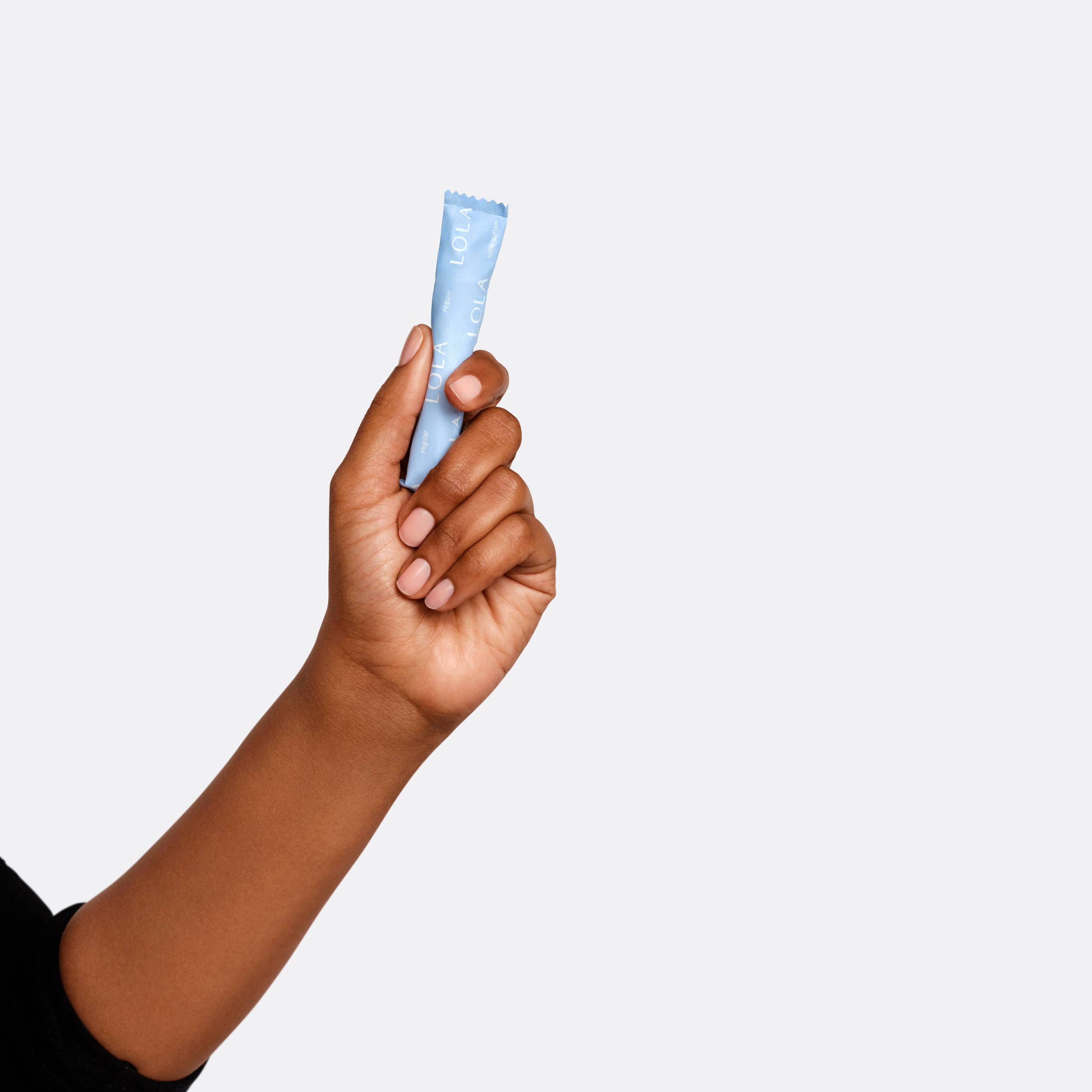 A hand with medium-brown skin holds up a small blue BPA-free Lola 20ct Regular Tampons applicator tube against a plain light background. The person is wearing a black long-sleeve shirt.