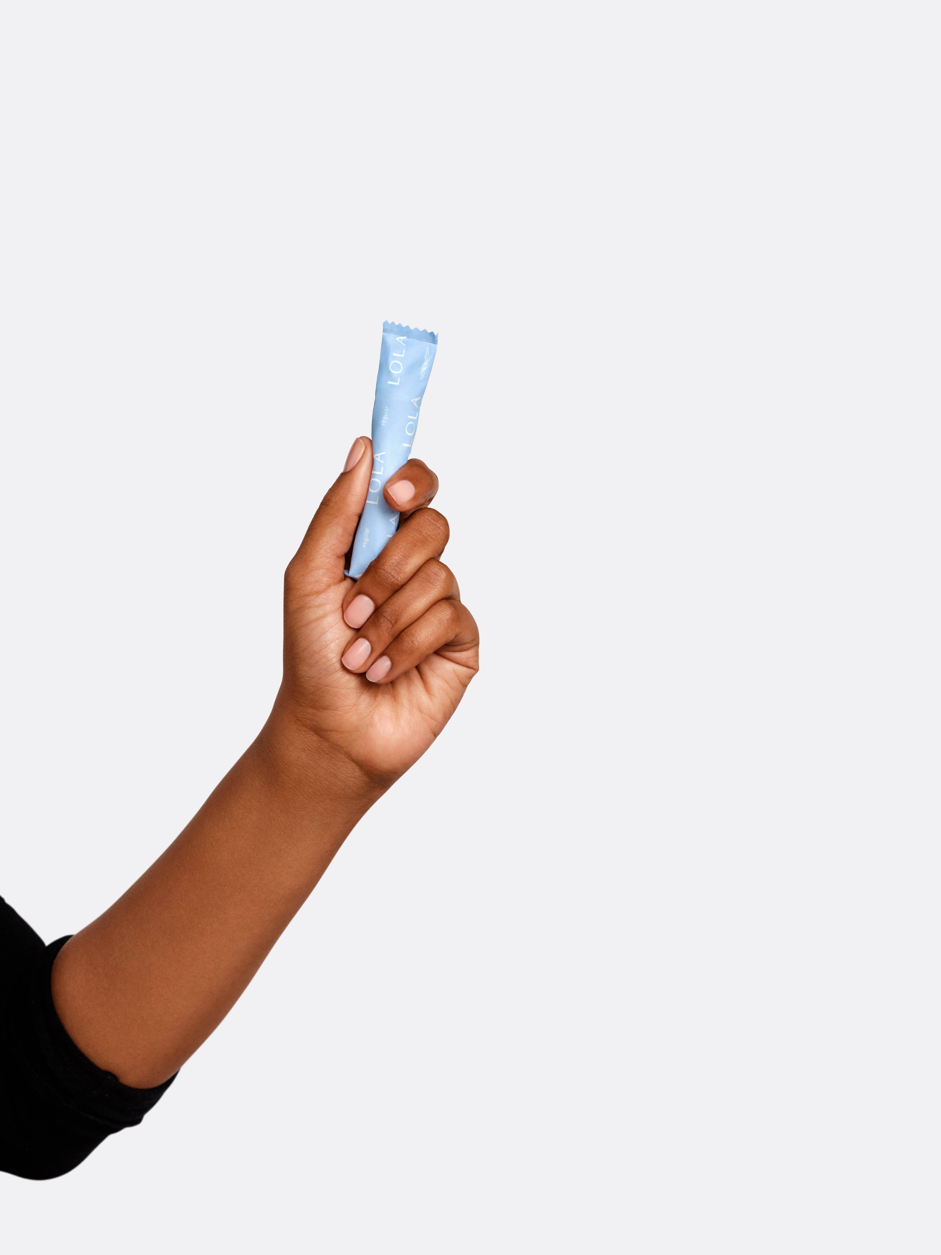 A hand with medium-brown skin holds up a small blue BPA-free Lola 20ct Regular Tampons applicator tube against a plain light background. The person is wearing a black long-sleeve shirt.