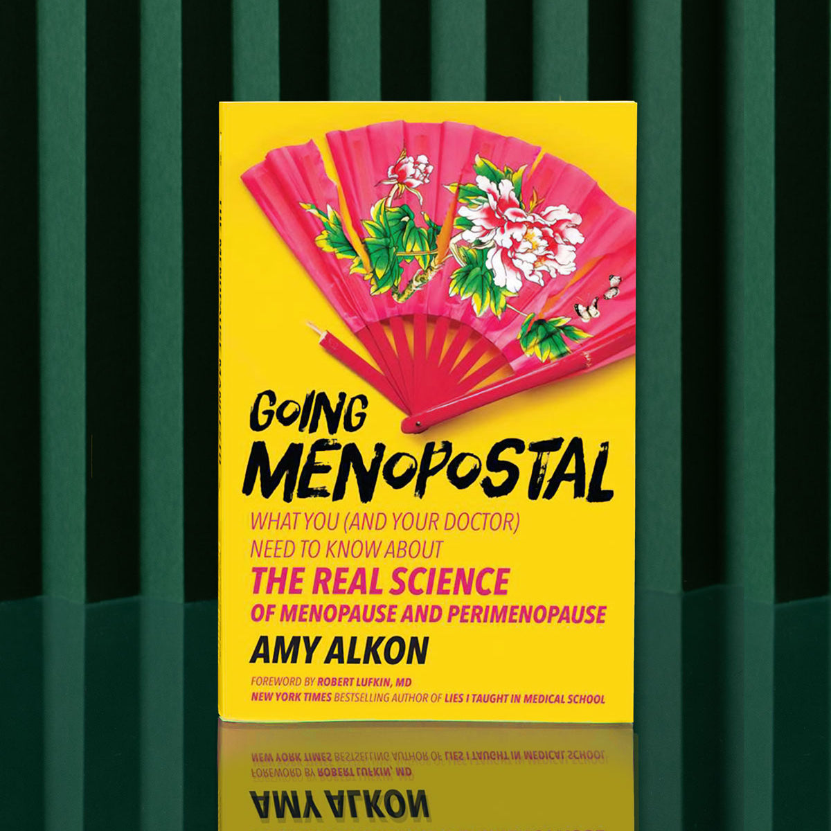 Going Menopostal by Amy Alkon is a yellow menopause guide standing upright on a green surface with dark green striped background; its cover shows a red fan decorated with pink and white flowers.
