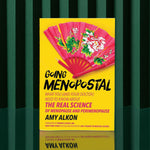 Going Menopostal by Amy Alkon is a yellow menopause guide standing upright on a green surface with dark green striped background; its cover shows a red fan decorated with pink and white flowers.