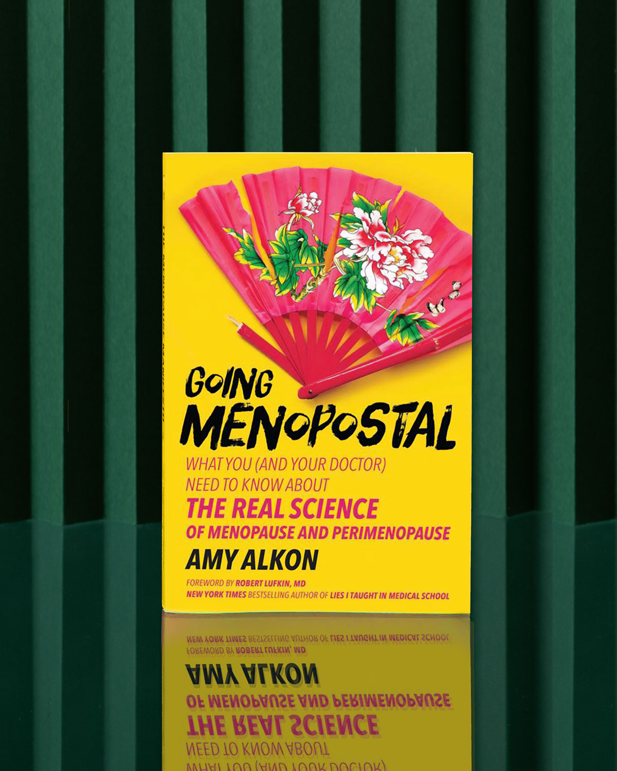 Going Menopostal by Amy Alkon is a yellow menopause guide standing upright on a green surface with dark green striped background; its cover shows a red fan decorated with pink and white flowers.
