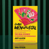Going Menopostal by Amy Alkon is a yellow menopause guide standing upright on a green surface with dark green striped background; its cover shows a red fan decorated with pink and white flowers.