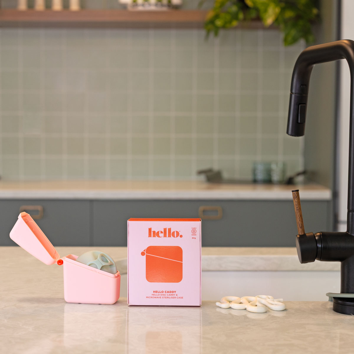 A Hello Caddy pink food-grade silicone container with a pacifier, white pacifier rings, and a box labeled "hello" sit on a kitchen counter with light green tiles, gray cabinets, and a sink in the background.