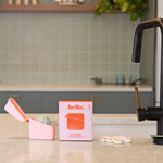 A Hello Caddy pink food-grade silicone container with a pacifier, white pacifier rings, and a box labeled "hello" sit on a kitchen counter with light green tiles, gray cabinets, and a sink in the background.