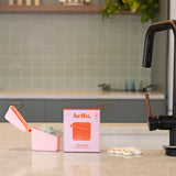 A Hello Caddy pink food-grade silicone container with a pacifier, white pacifier rings, and a box labeled "hello" sit on a kitchen counter with light green tiles, gray cabinets, and a sink in the background.