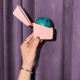 A hand with metallic nail polish holds a small pink Hello Caddy with a teal food-grade silicone item inside, set against a purple curtain background.