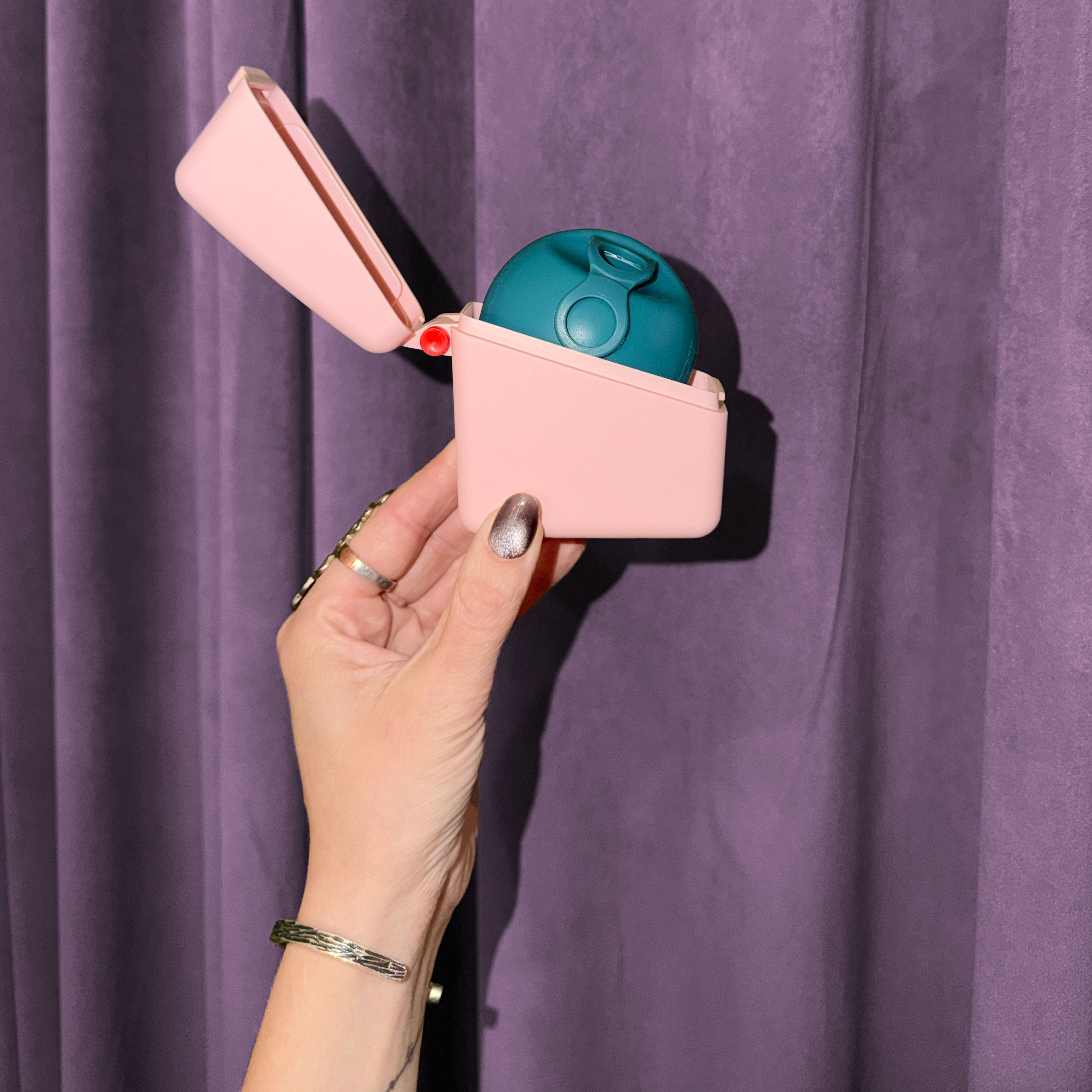 A hand with metallic nail polish holds a small pink Hello Caddy with a teal food-grade silicone item inside, set against a purple curtain background.