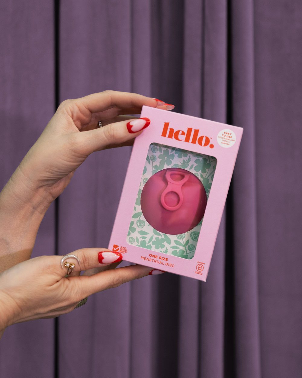 A person with red and white manicured nails holds the Hello Disc - Bubblegum box, featuring an easy-removal, leak-proof menstrual disc, set against a floral backdrop and purple curtain.