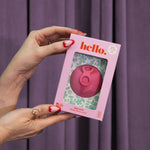 A person with red and white manicured nails holds the Hello Disc - Bubblegum box, featuring an easy-removal, leak-proof menstrual disc, set against a floral backdrop and purple curtain.