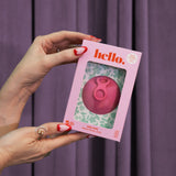 A person with red and white manicured nails holds the Hello Disc - Bubblegum box, featuring an easy-removal, leak-proof menstrual disc, set against a floral backdrop and purple curtain.