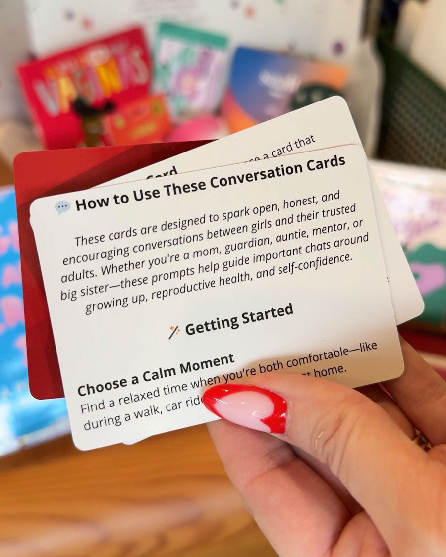 A hand with red and white nail art holds a card titled “How to Use These Conversation Cards” from Red Tribe Period Conversation Cards, designed to spark important puberty talks and discussions about growing up and self-confidence. Blurred cards appear in the background.
