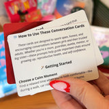 A hand with red and white nail art holds a card titled “How to Use These Conversation Cards” from Red Tribe Period Conversation Cards, designed to spark important puberty talks and discussions about growing up and self-confidence. Blurred cards appear in the background.