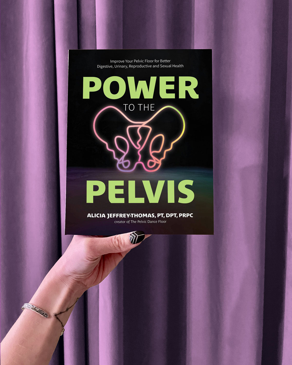 A hand with painted nails holds Power to the Pelvis against a purple curtain. The book cover features bold green and white text and a neon outline of a pelvis, emphasizing pelvic floor physical therapy.