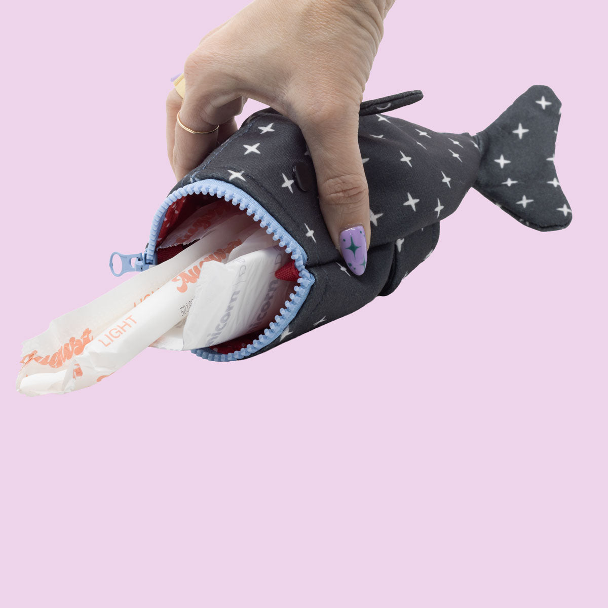 A hand holds the Shark Week Pouch, its zipper mouth partly open to show several period products inside. The person’s purple nail art pops against the playful light pink background.