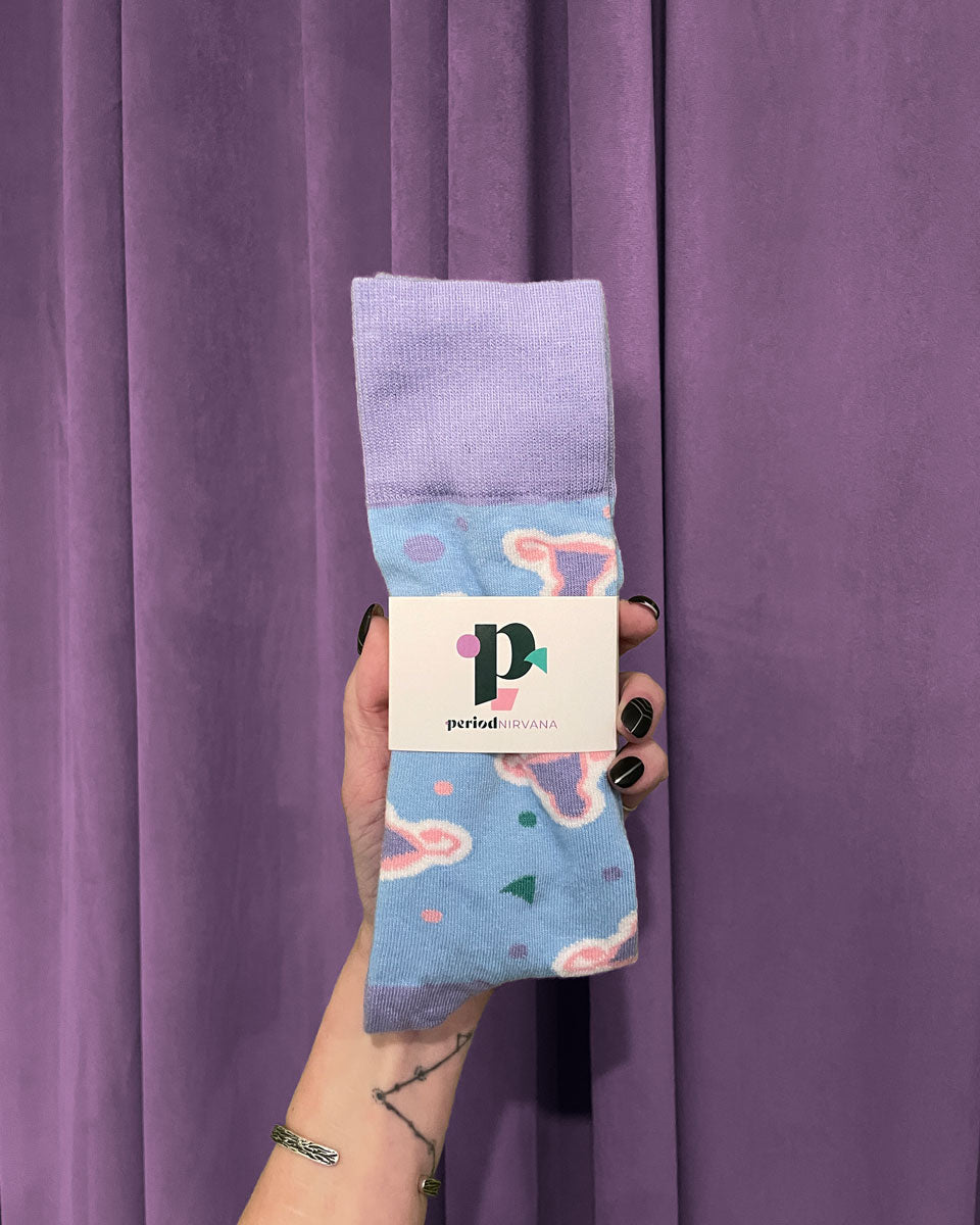 A hand with black nail polish holds Period Nirvana Cotton Uterus Socks—pastel blue crew socks featuring pink and green abstract shapes, made from premium combed cotton—against a purple curtain, with a Period Nirvana business card tucked into the socks.