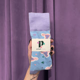 A hand with black nail polish holds Period Nirvana Cotton Uterus Socks—pastel blue crew socks featuring pink and green abstract shapes, made from premium combed cotton—against a purple curtain, with a Period Nirvana business card tucked into the socks.