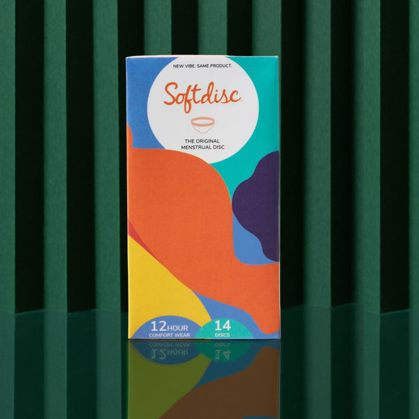 A vibrant box of Softdisc, featuring 14 disposable menstrual discs, stands upright on a green surface with a dark green striped background. The packaging highlights leak-proof period protection and up to 12 hours of comfortable wear.
