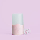 The Hello Steamy Bundle, featuring a pink and white cylindrical menstrual cup sanitizer with "hello" embossed on the front and a small clear measuring cup with red markings, is displayed against a light pink background.