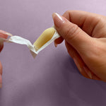 A person with manicured nails peels open a foil wrapper to reveal a NeuEve Velvet 70+ Suppository, designed to support vaginal health, against a light purple background.